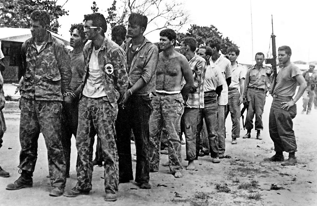 Captured members of Brigade 2506 after the failed Bay of Pigs invasion, April 1961 — a line of dishevelled men in camouflage fatigues guarded by a Cuban soldier with a rifle