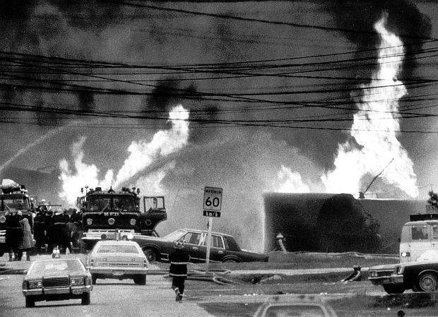 Street-level view of multiple propane fires burning at the derailment site, with fire trucks, police cars, and a 60 km/h speed sign visible in the foreground